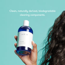 Hand holding a blue cleaning bottle with text about natural and biodegradable components on a teal background.