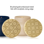 Two round brushed-gold embossed metal candle lid coasters with a sculpted wavy edge, displayed in front of a lit cream candle and a dark blue container on a white background, with text reading 'Brushed-gold embossed metal lids with sculpted, wavy edge'.