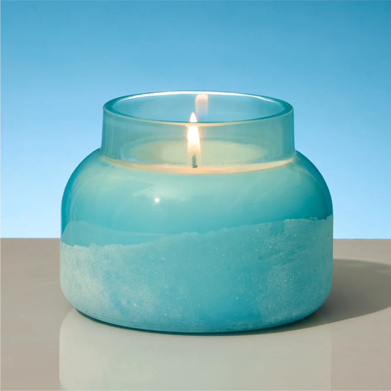 A lit teal glass jar candle with a flared rim sits on a light gray surface against a blue gradient background.