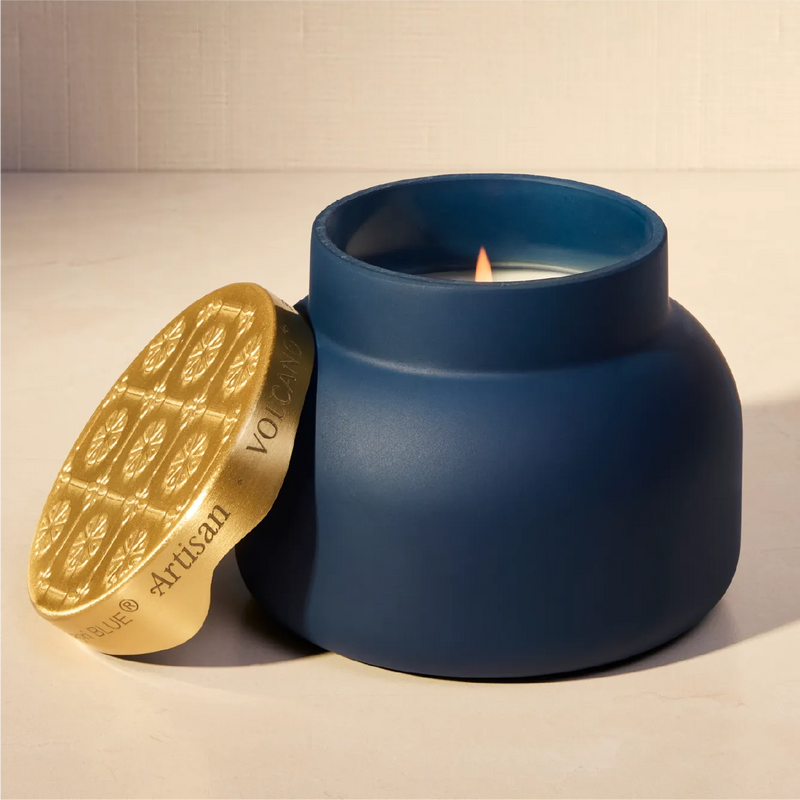 A matte navy blue jar candle with a lit wick, shown with its matching metallic gold lid leaning against it on a light beige surface.