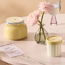 Two candles with striped lids on a marble surface, next to a vase with pink flowers and a glass of water.