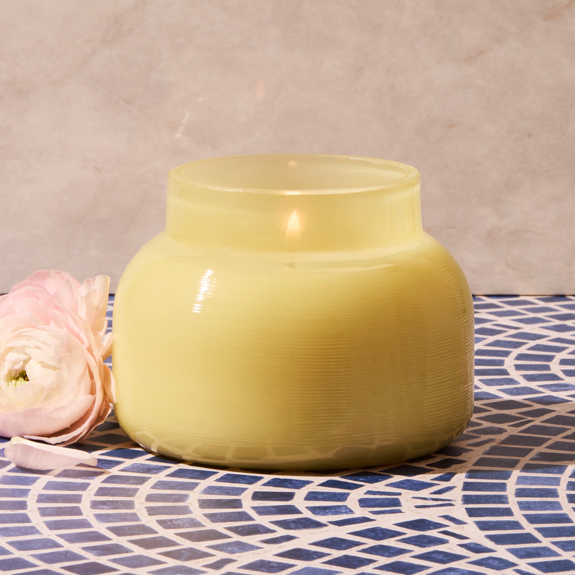 Yellow candle in a textured jar on a patterned surface with a pink flower.