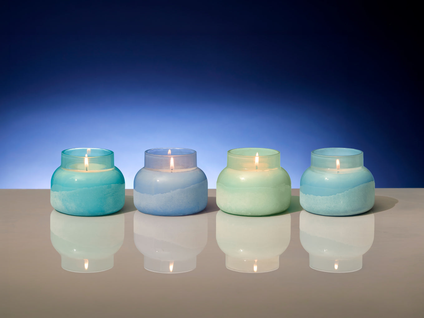Four lit candles in gradient blue glass holders on a reflective surface with a blue background