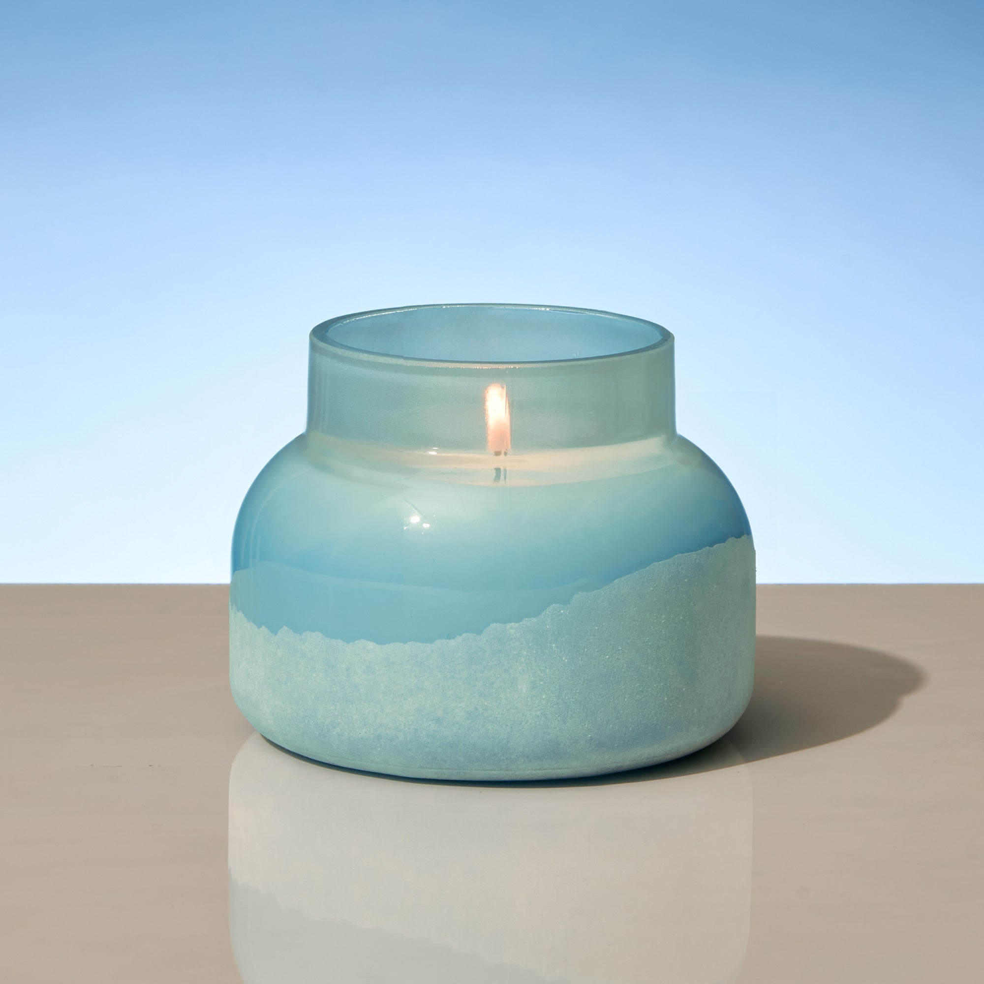 Candle in a blue glass jar on a reflective surface with a light blue background