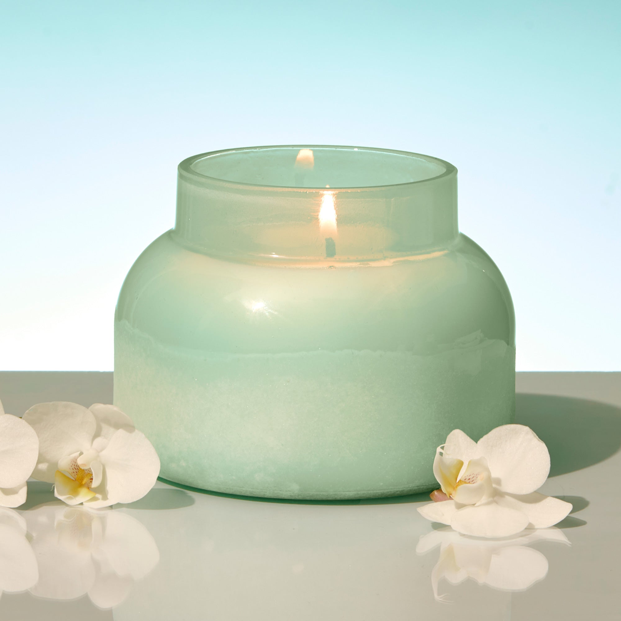 Green candle in a glass jar with white flowers on a reflective surface