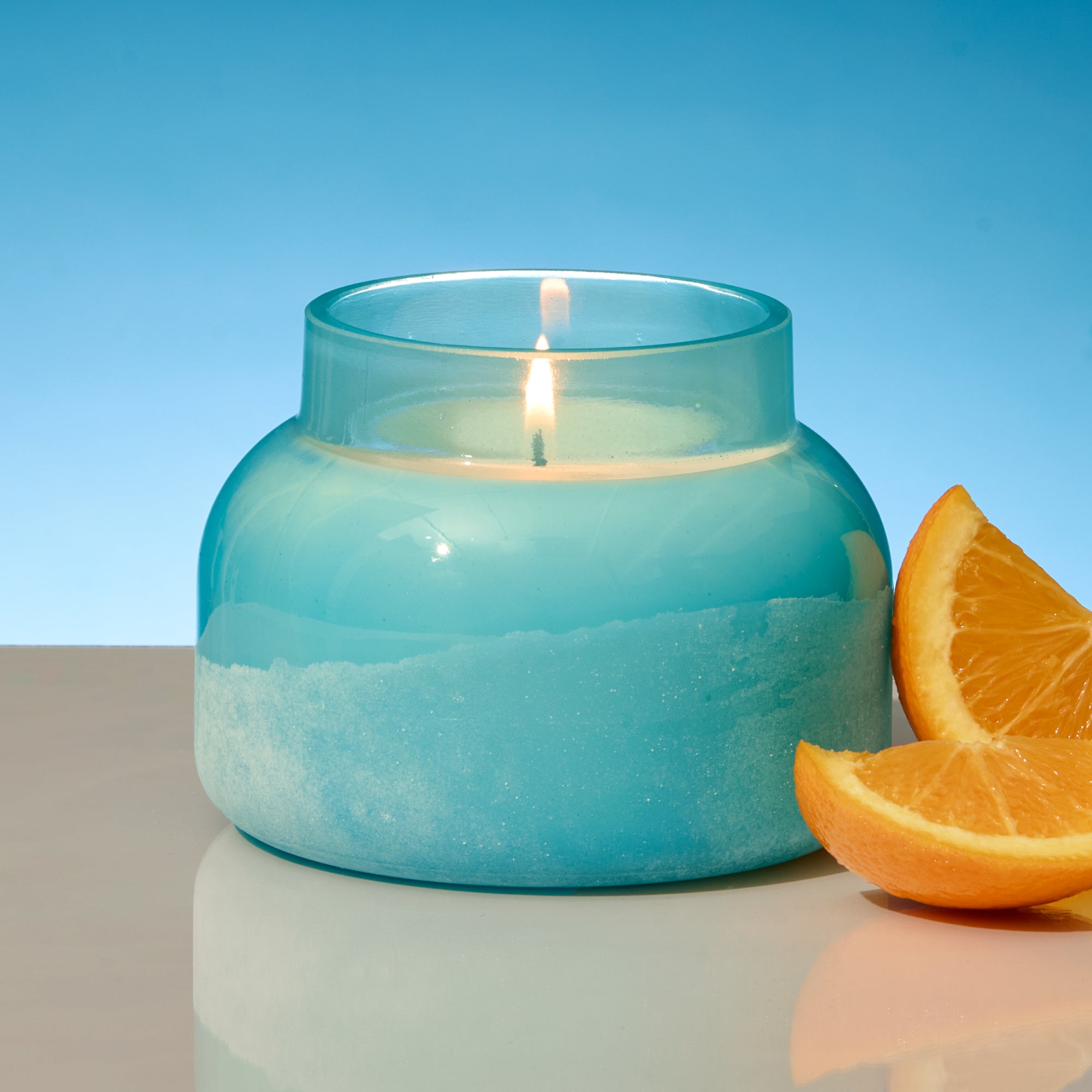 Blue candle in a glass jar with a slice of orange on a reflective surface and blue background