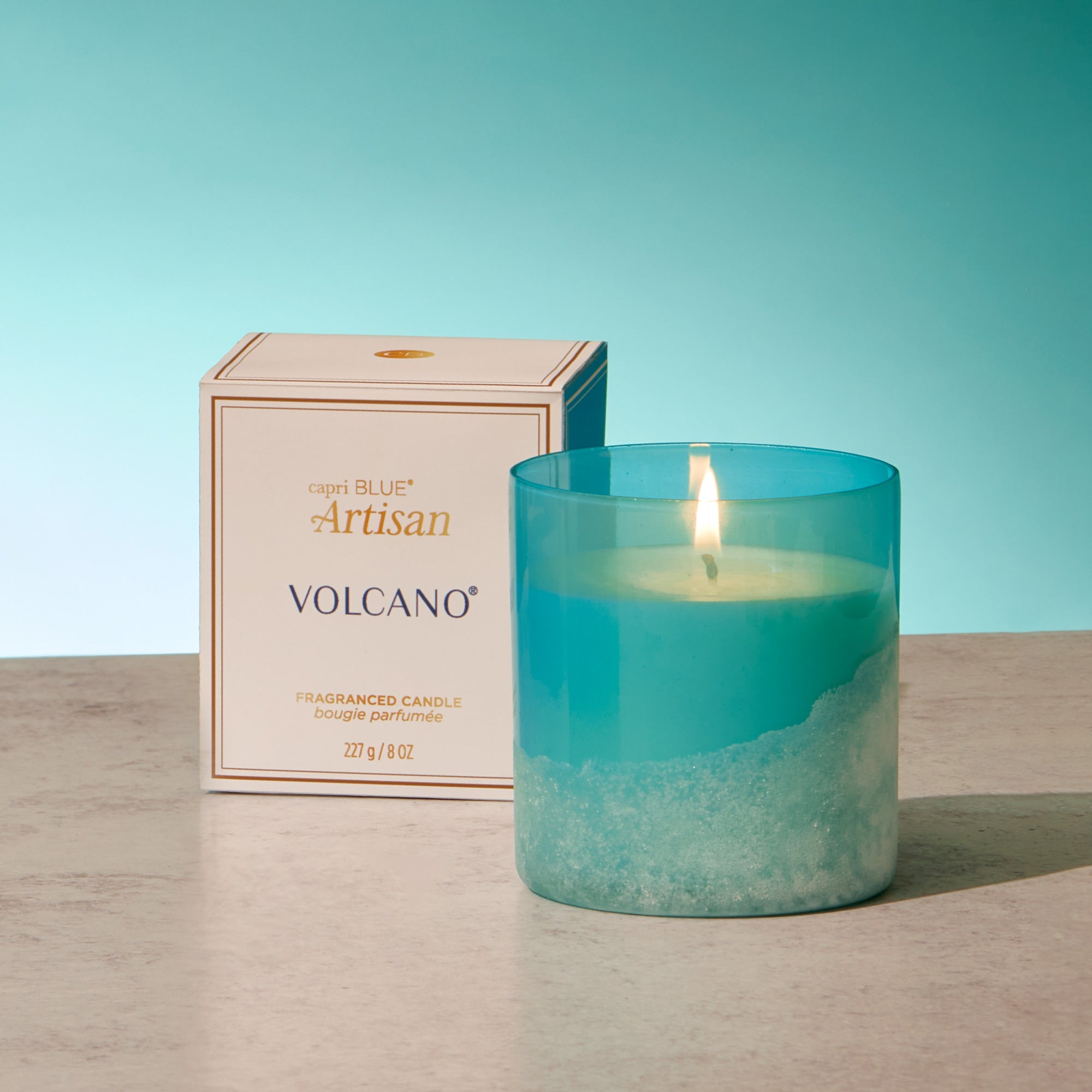 Teal candle with a lit wick next to its packaging on a wooden surface with a teal background