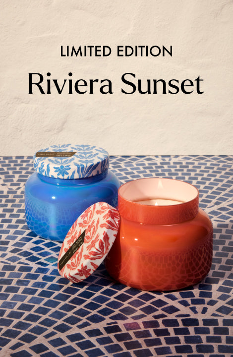 Two candles, one blue and one red, with floral lids on a textured surface, labeled 'Limited Edition Riviera Sunset'.