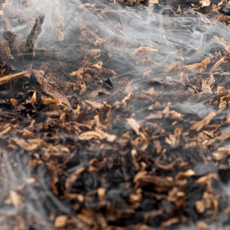 Close-up of loose tobacco leaves in shades of dark brown, tan, and black, with visible smoke rising through the leaves.