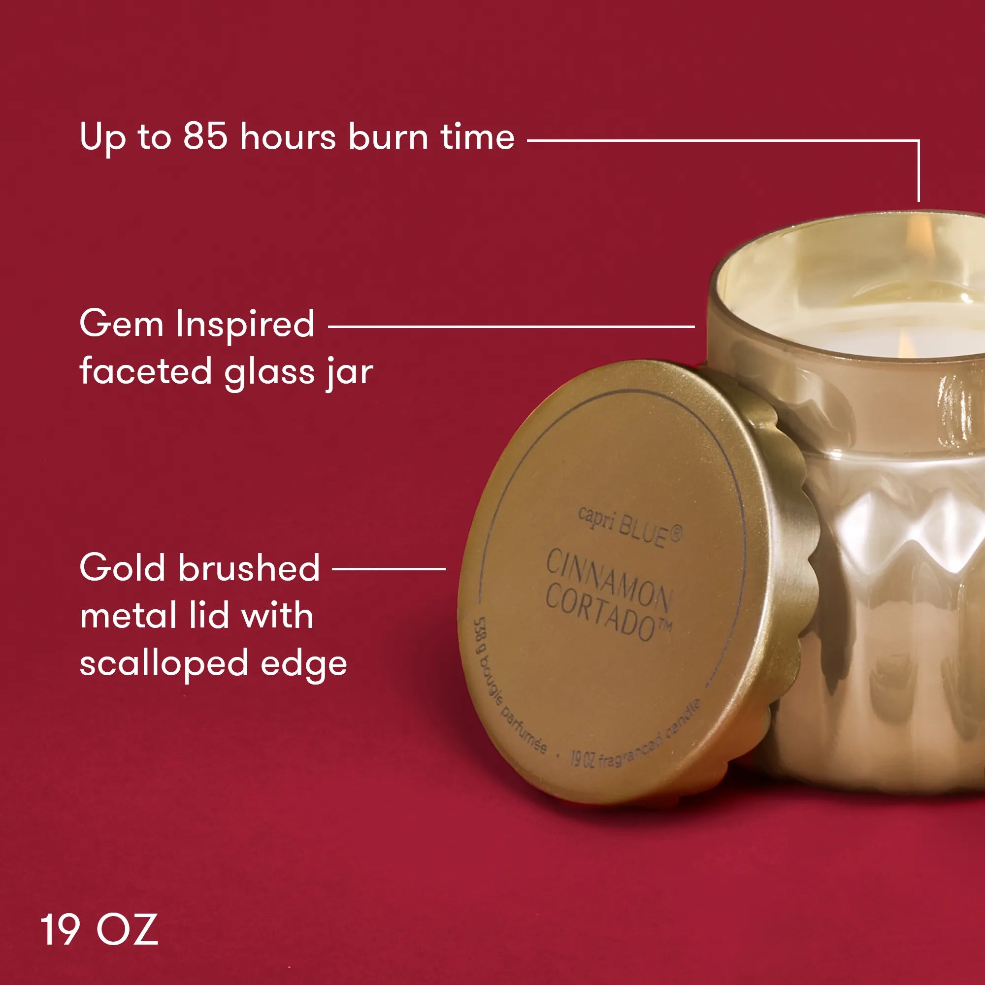 A lit, Cinnamon Cortado Royal Gem Signature Candle in a champagne-topaz  faceted glass jar wit text describing its features:  "Up to 85 hours burn time", "Gem inspired faceted glass jar", "Gold brushed metai lid with scalloped edge" and "19 oz."