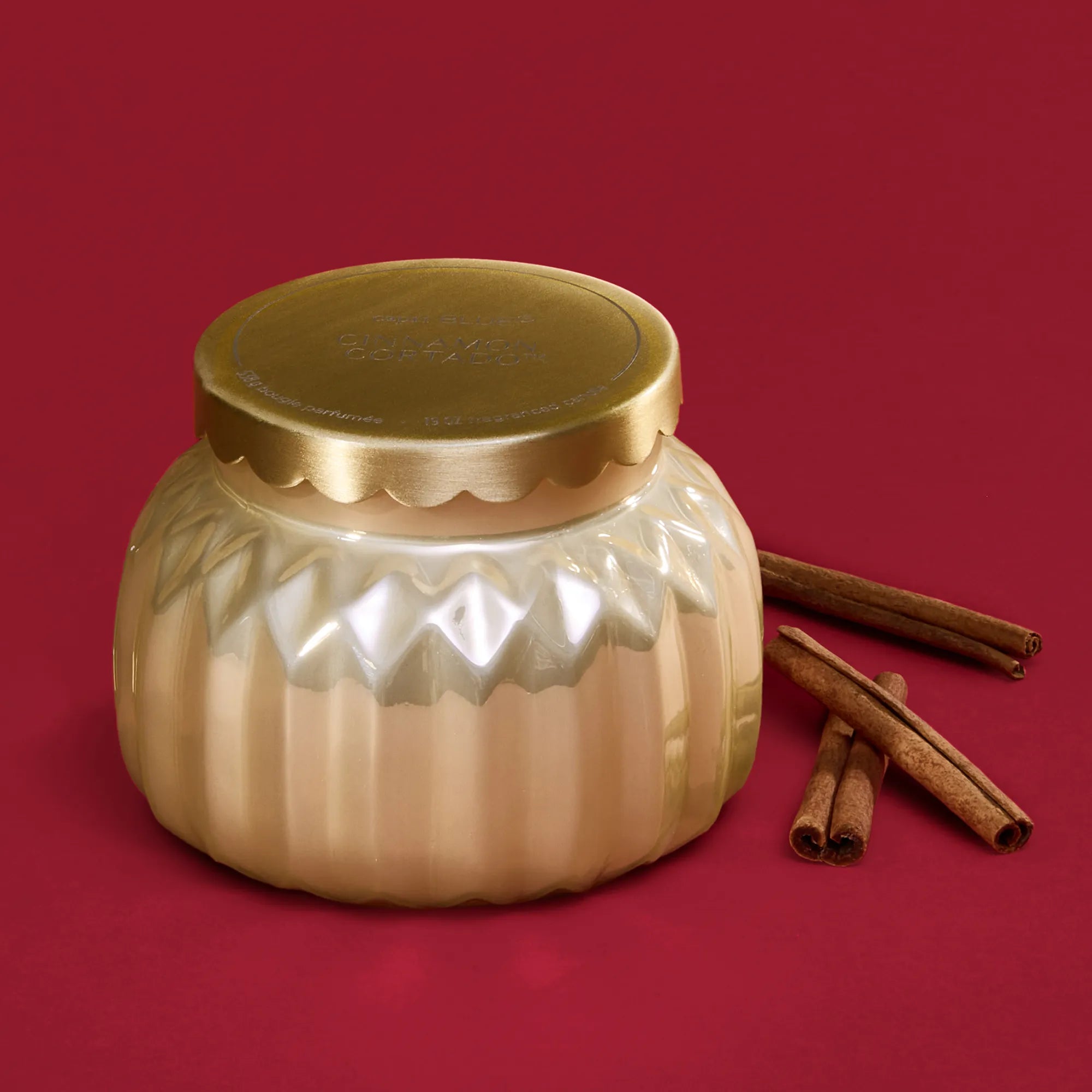 A Cinnamon Cortado Royal Gem Signature Candle in a champagne-topaz, faceted glass jar  with a scalloped gold lid beside cinnamon sticks.