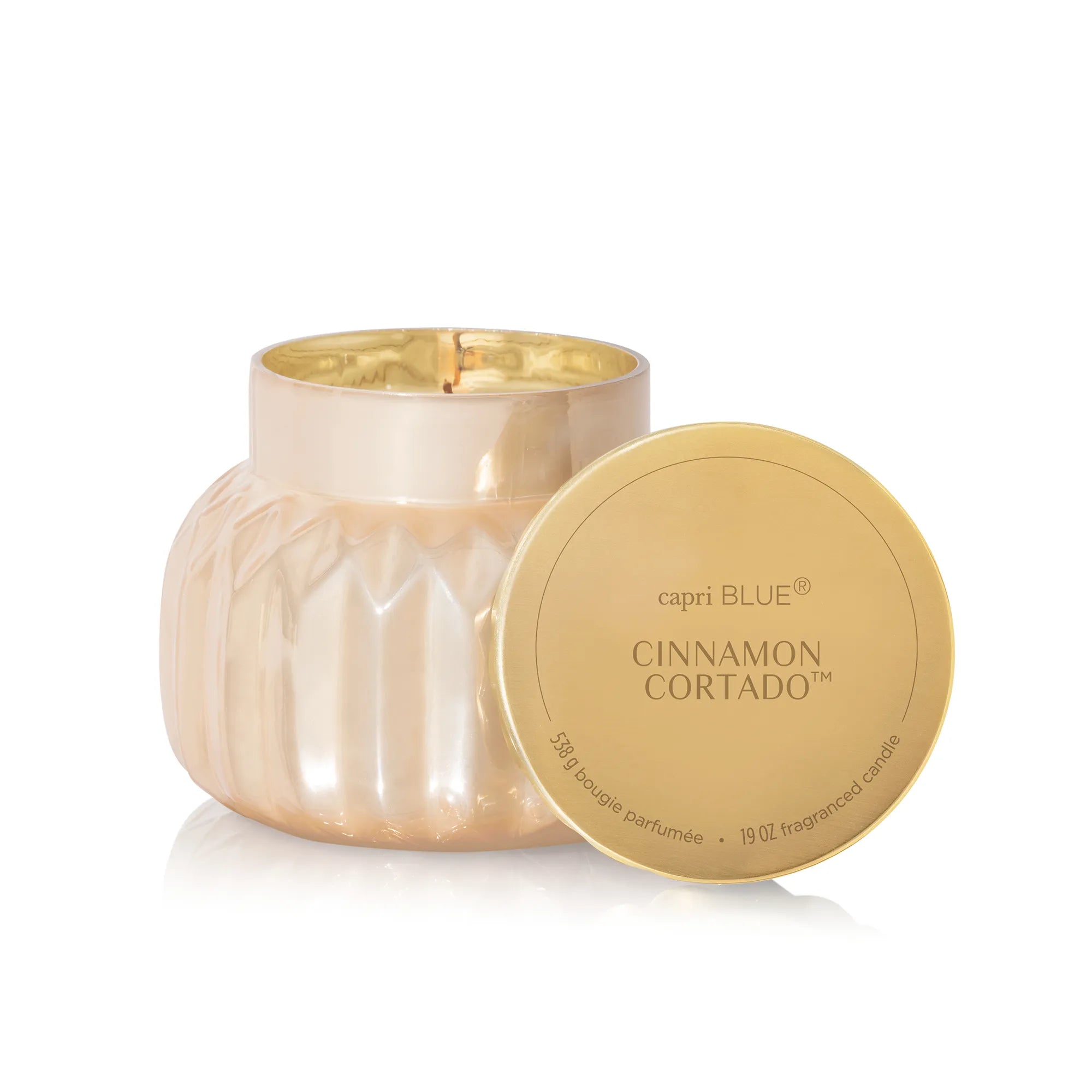 A lit, Cinnamon Cortado Royal Gem Signature Candle in a champagne-topaz, faceted glass jar beside its scalloped gold lid.