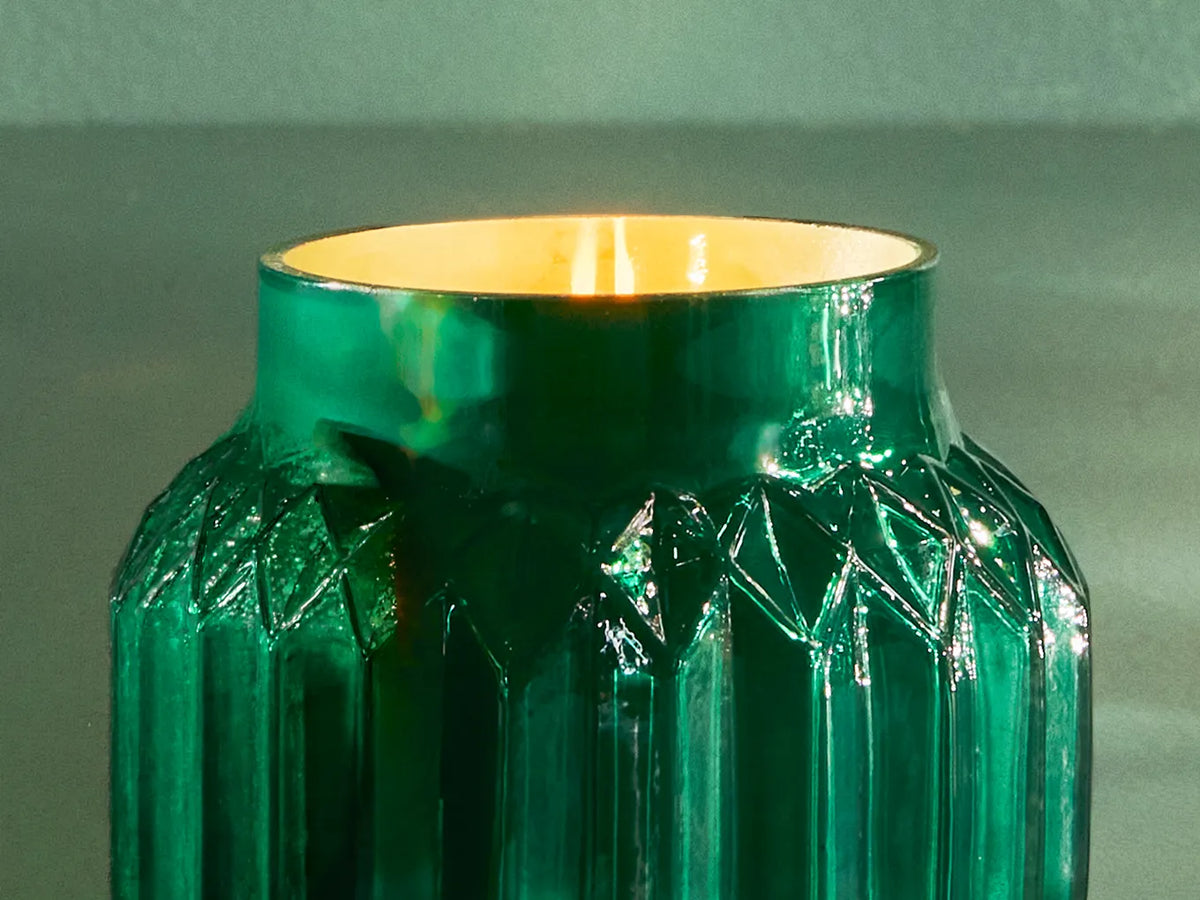 A lit, Volcano Royal Gem Petite Candle with an emerald green, gem-inspired jar.