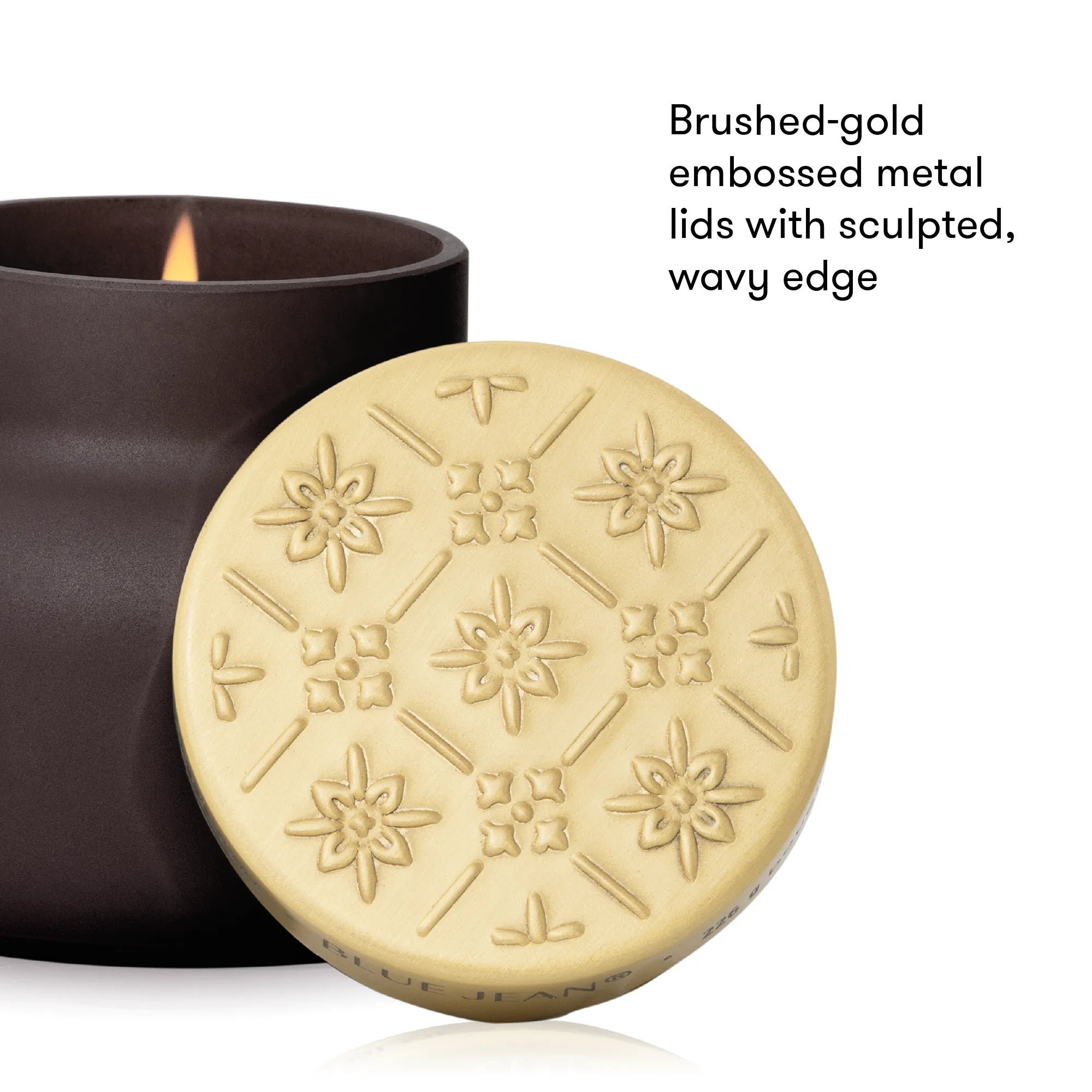 A close-up product shot showing a round brushed-gold metal candle lid with embossed floral and geometric patterns, placed in front of a dark brown candle jar with a visible flame. Text on the right reads, 'Brushed-gold embossed metal lids with sculpted, wavy edge.