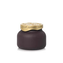 A single jar of Blue Jean Artisan spread on a clean white background. The jar has a dark, matte purple-brown body and a metallic gold lid with the brand name 'Capri Blue Artisan' printed on it.