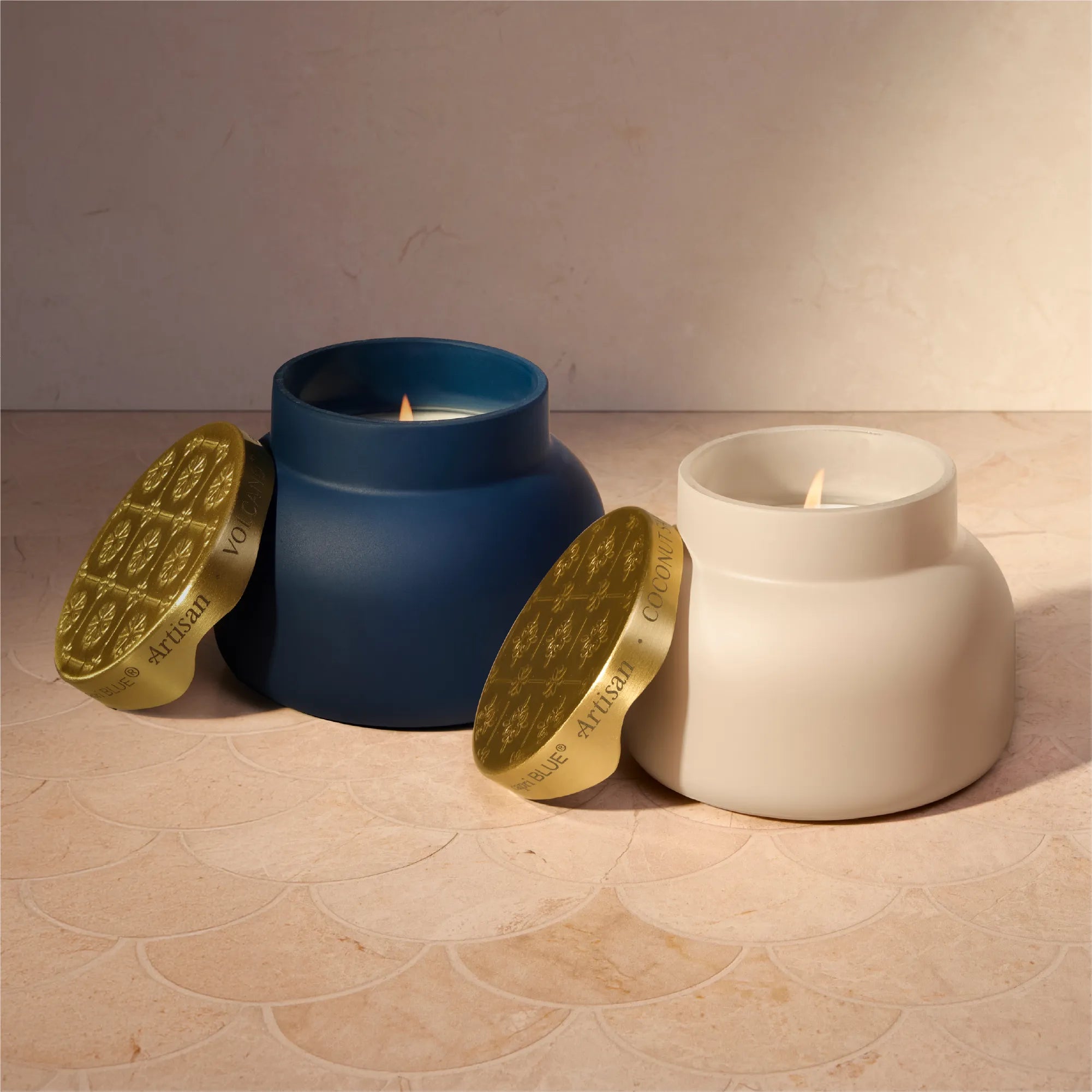 Two ceramic candles in matte blue and off-white pots with gold lids, shown lit on a beige tiled surface against a beige wall.