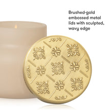 A round brushed-gold metal candle lid with embossed floral and geometric patterns, shown in front of a cream-colored candle. Text on the right reads, 'Brushed-gold embossed metal lids with sculpted, wavy edge.