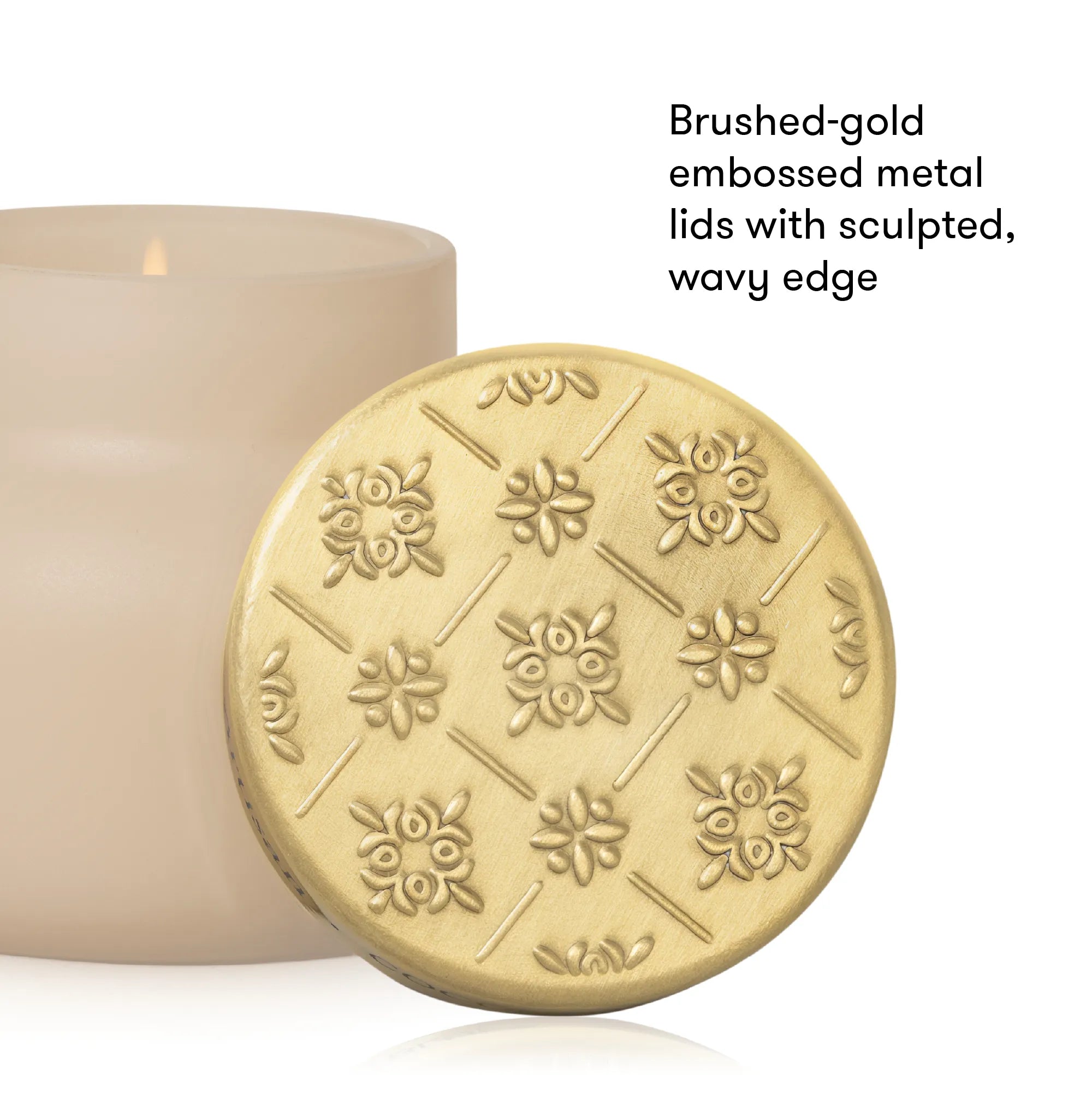 A round brushed-gold metal candle lid with embossed floral and geometric patterns, shown in front of a cream-colored candle. Text on the right reads, 'Brushed-gold embossed metal lids with sculpted, wavy edge.