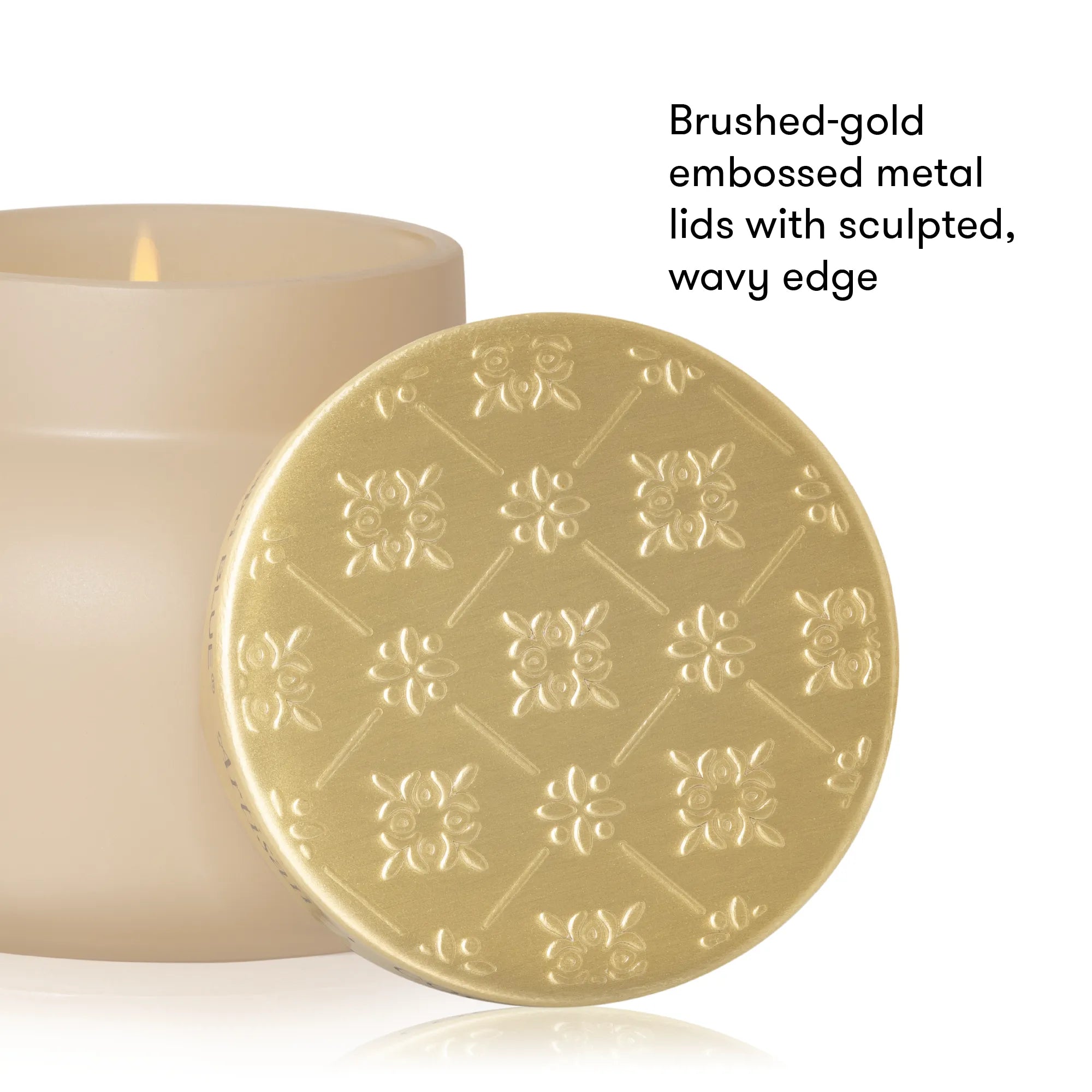 A close-up product shot showing a round brushed-gold metal candle lid with embossed floral patterns and a wavy edge, placed in front of a cream-colored candle. Black text on the right reads, 'Brushed-gold embossed metal lids with sculpted, wavy edge.