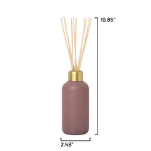 A reed diffuser bottle in a muted mauve-pink color with a gold collar and natural beige reeds, shown on a white background with black dimension lines indicating height of 10.85 inches and width of 2.