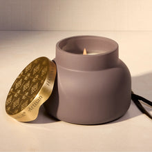 A matte taupe-gray ceramic candle jar with a lit flame, shown next to its gold-toned lid engraved with floral motifs and the brand name 'HAVANA VANILLA'.
