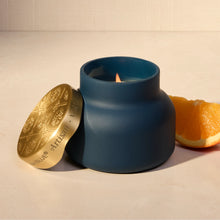 A matte navy blue candle jar with a small flame, shown with its gold lid leaning beside it on a light surface. A slice of orange is placed behind the candle.