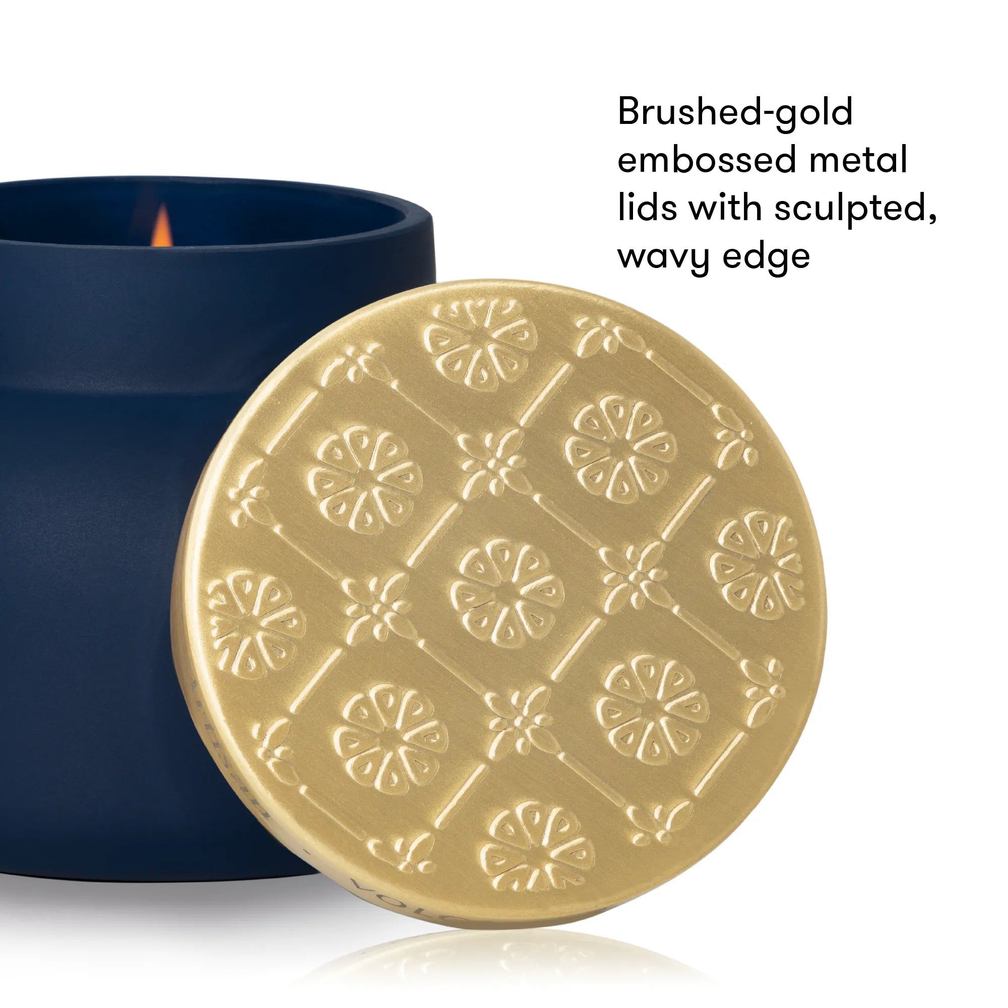 A brushed gold metal lid with an embossed floral pattern and wavy edge, shown leaning against a dark navy blue candle jar on a white background. Black text on the right reads: 'Brushed-gold embossed metal lids with sculpted, wavy edge.
