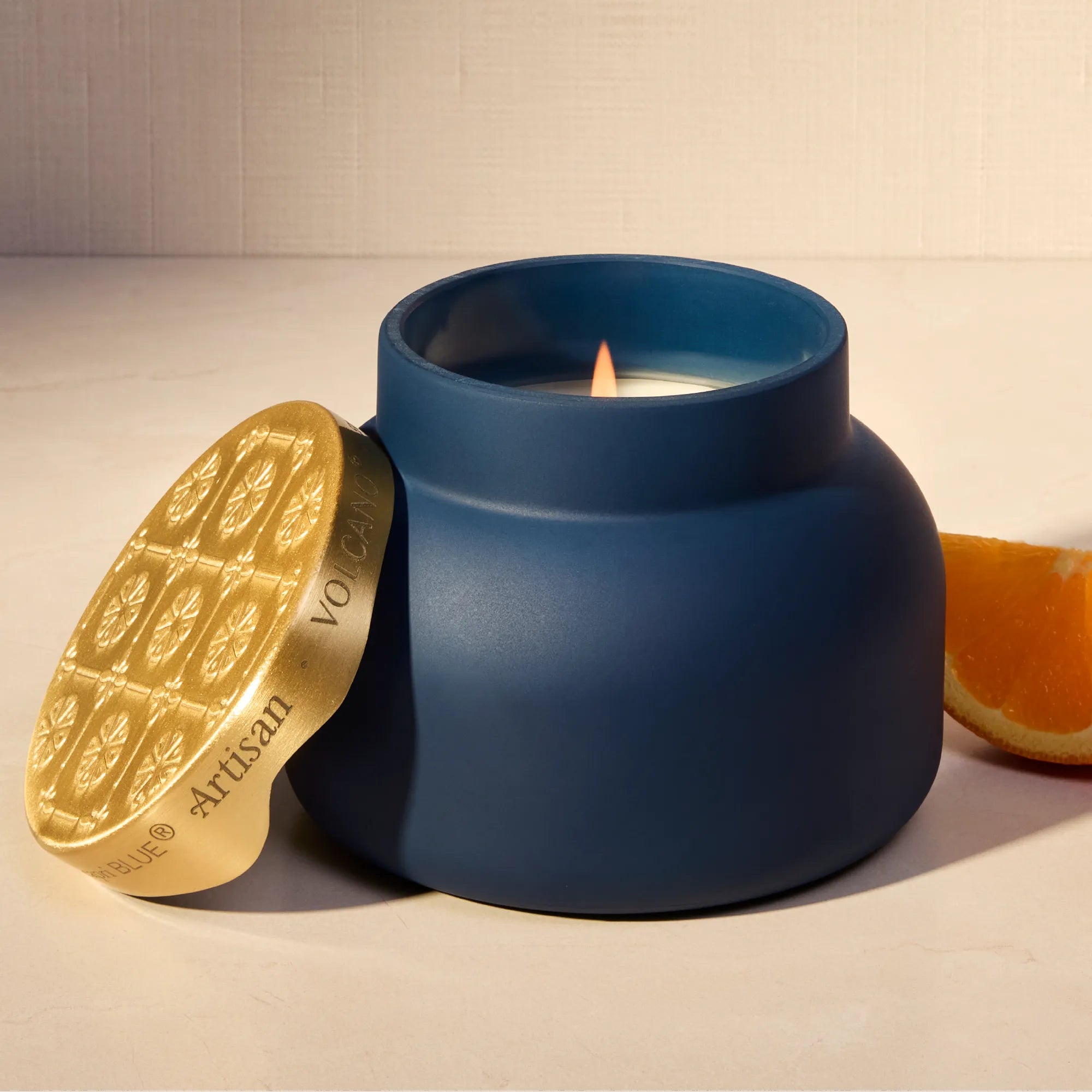 A matte navy blue jar candle with a gold lid leaning against it on a light beige surface. A lit candle flame is visible inside the jar, and a slice of orange sits to the right.