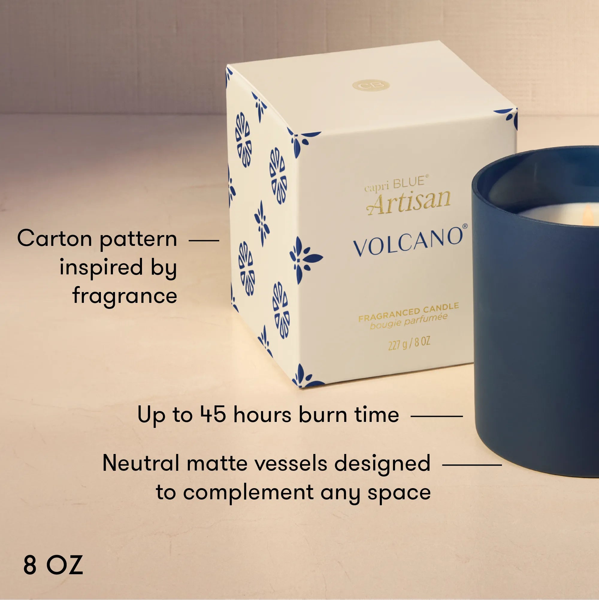 A Volcano Artisan scented candle by Capri Blue. The product features a white square box with blue floral patterns and gold lettering, next to a matte navy blue cylindrical vessel.