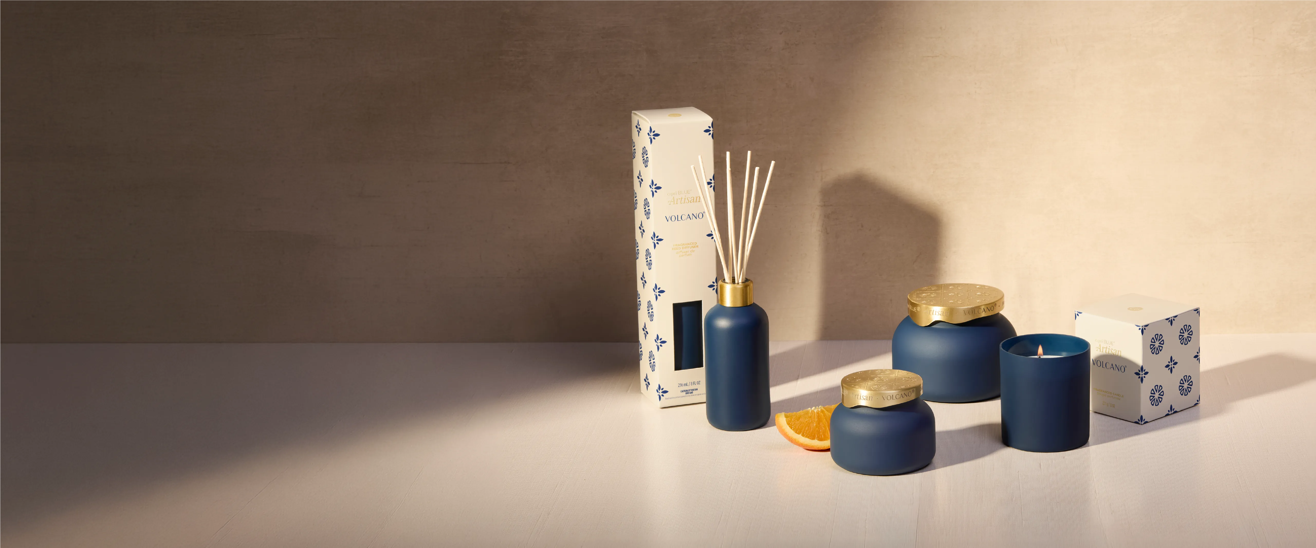 A luxury home fragrance set featuring a reed diffuser, three scented candles in matte navy blue containers with gold lids, and two patterned boxes, styled on a light beige surface against a textured beige wall.