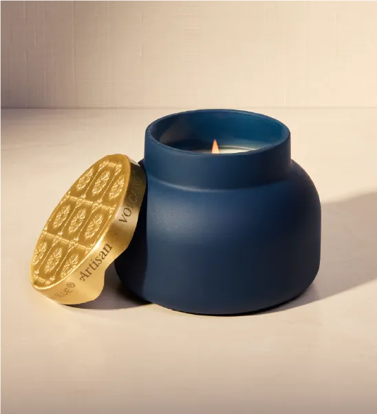 A matte navy blue jar candle with a lit flame, shown next to its gold lid which features a chocolate bar design and the brand name 'VOLCANO'.