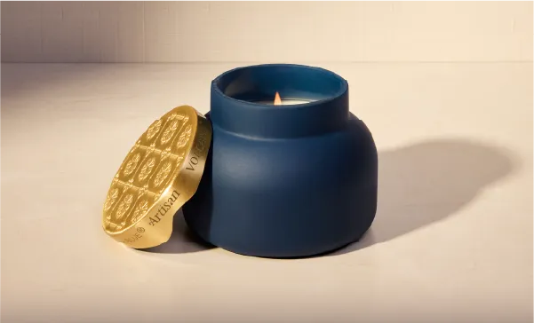 A matte navy blue jar candle with a lid shaped like a gold chocolate bar, shown on a warm beige background with the lid leaning against the jar.