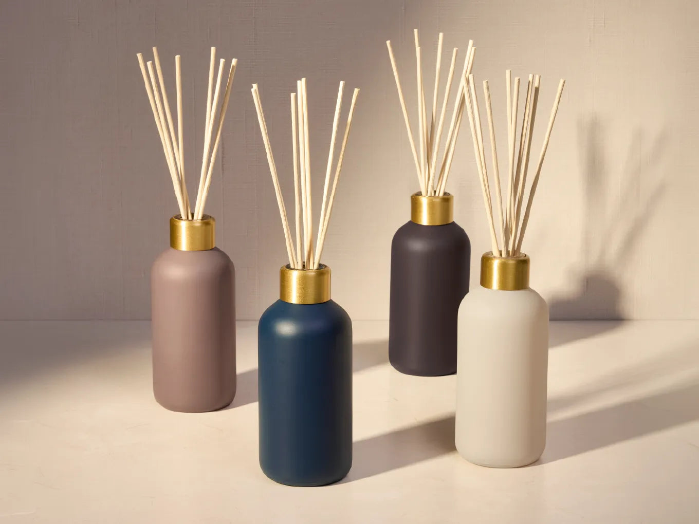 Four matte reed diffusers in muted neutral colors with natural reed sticks, arranged on a light surface against a warm beige background.