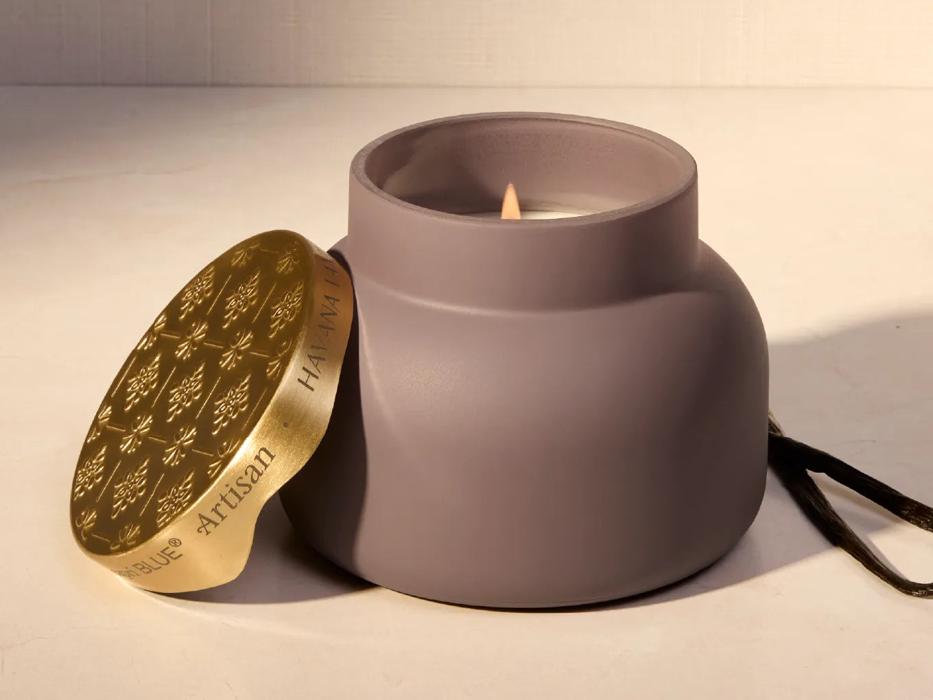 A matte taupe-gray ceramic candle jar with a lit flame, shown next to its gold-toned lid engraved with floral motifs and the words 'Artisan' and 'HAVANA VANILLA'.