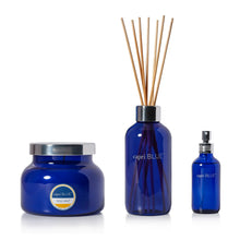Three blue scented products on a white background: a round jar candle with a silver lid and a round reed diffuser bottle with wooden reeds, plus a small atomizer spray bottle. The labels read 'capri BLUE' and 'VOLCANO'.