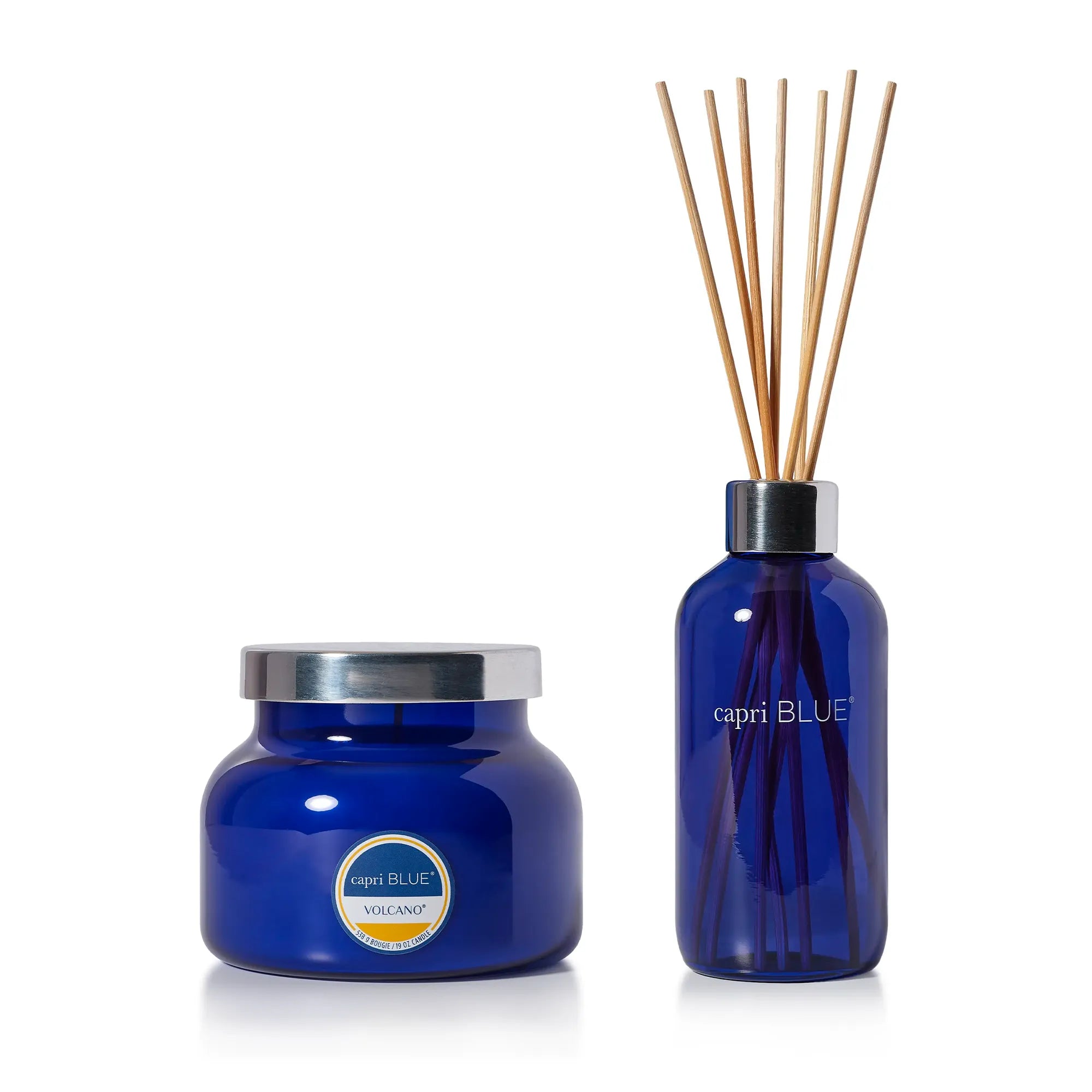 A blue glass reed diffuser set with a matching jar and several natural wood reeds, on a clean white background. The jar features a silver lid and a circular label that reads 'capri BLUE' and 'VOLCANO'.