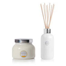 A reed diffuser set with a white jar candle and a white reed diffuser bottle with natural wood reeds, both featuring silver caps and a round label that reads 'CAPRI BLUE VOLCANO'.