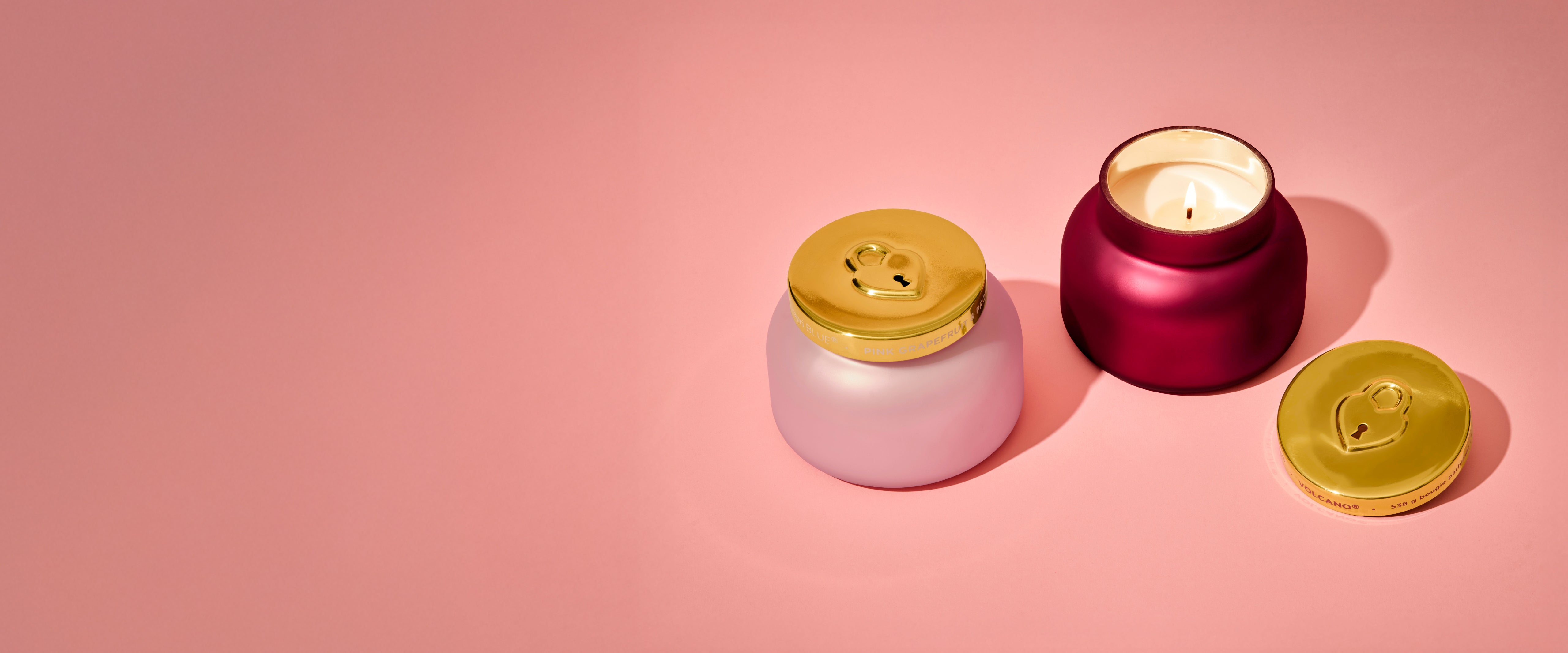 Two Locked In Love candles with gold lids on a pink background