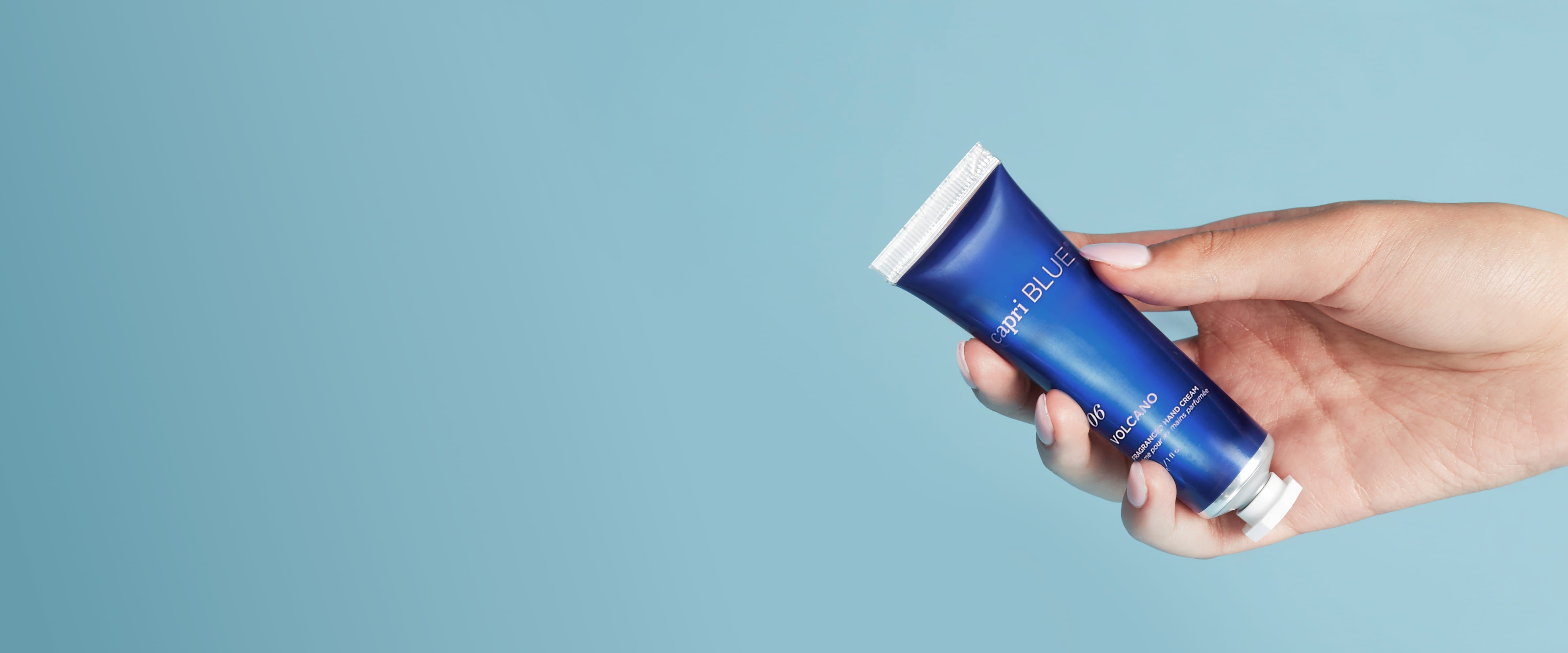 Hand holding a capri BLUE Volcano Mini Hand Cream on a blue background