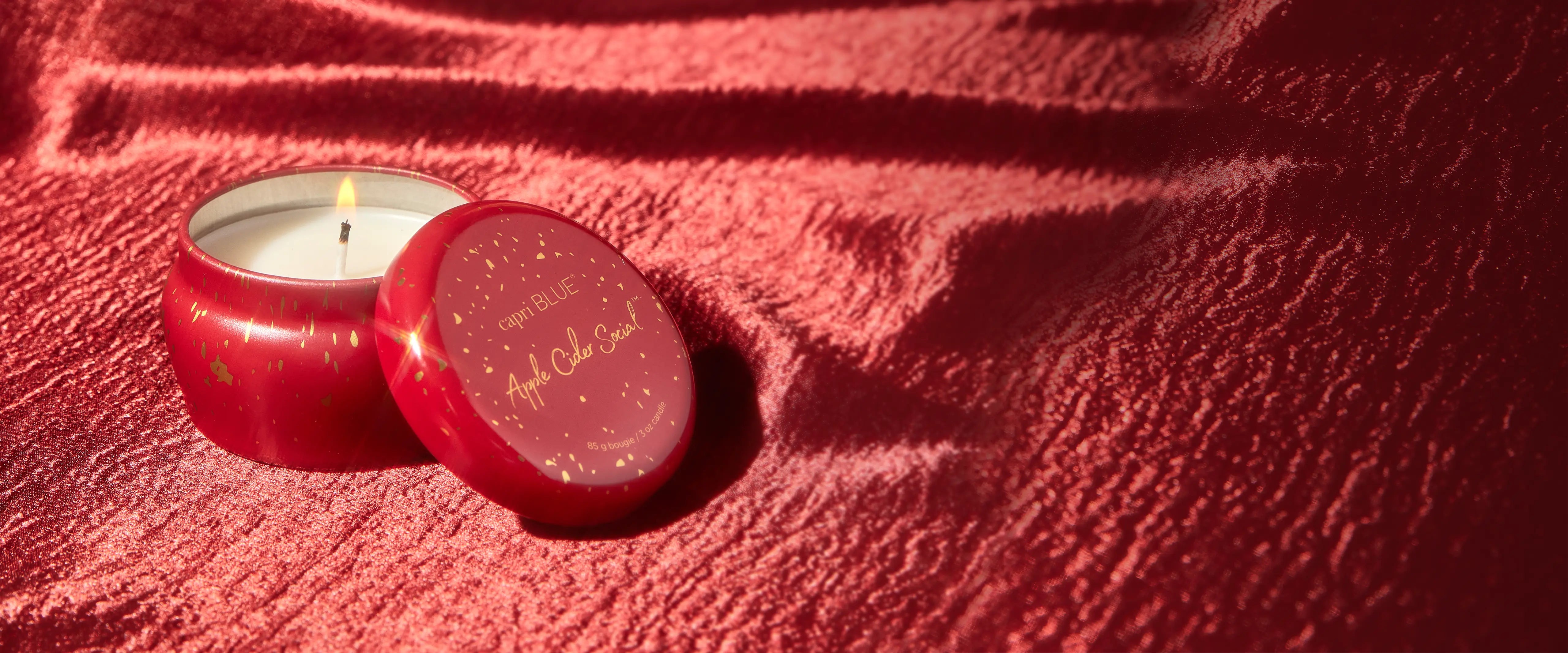 An open and lit Apple Cider Social Mini Tin Candle on a textured, red fabric.  Its lid leaning against it.