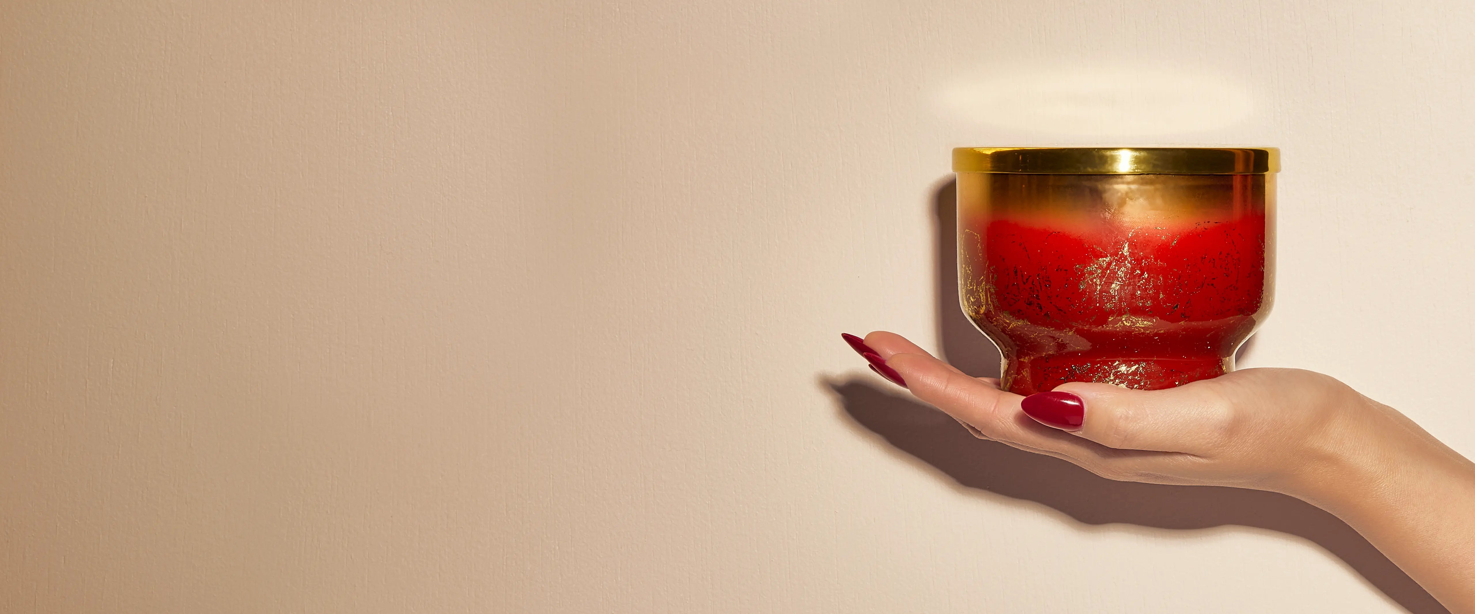 A lidded, red and gold Tinsel & Spice Glimmer Inverted Candle resting in an outstretched hand with red manicured fingernails.