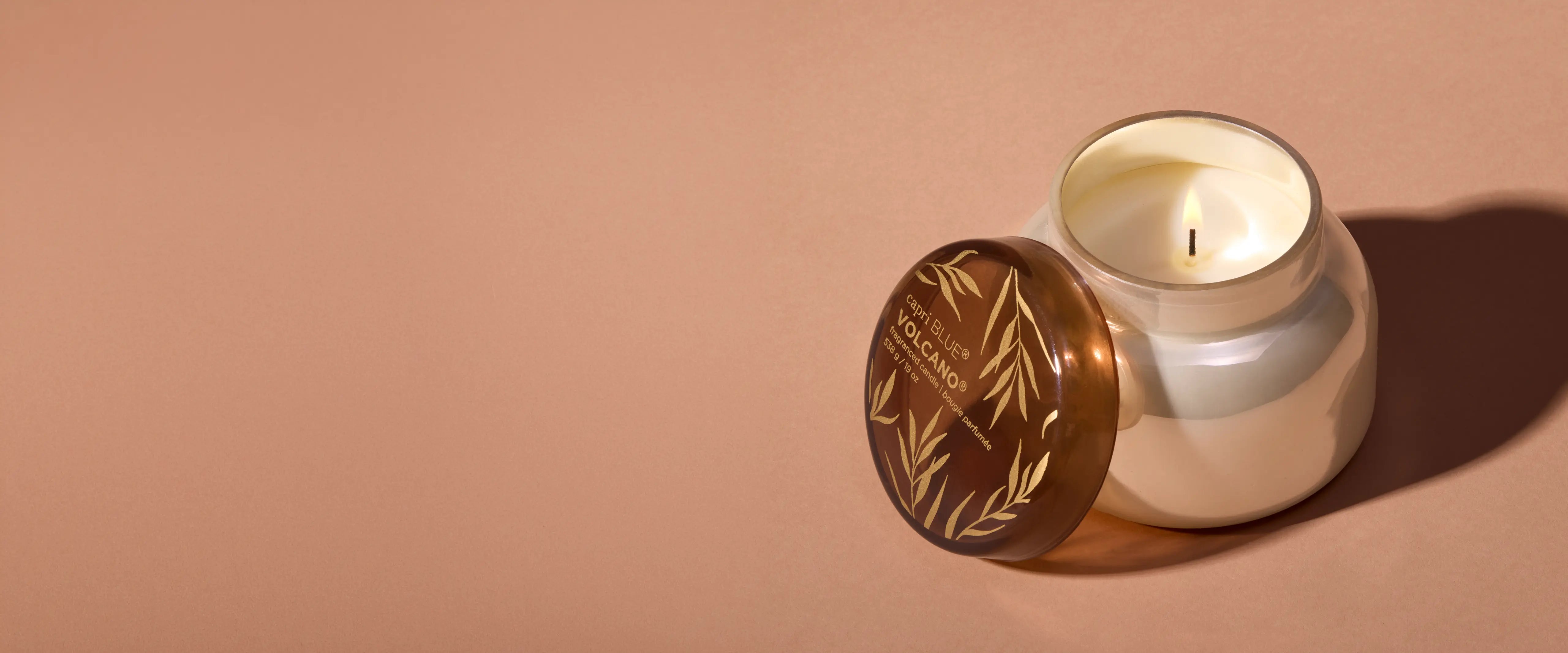A lit and uncovered Autumn Glow Volcano candle, its mocha brown lid resting against its pearlescent jar. 