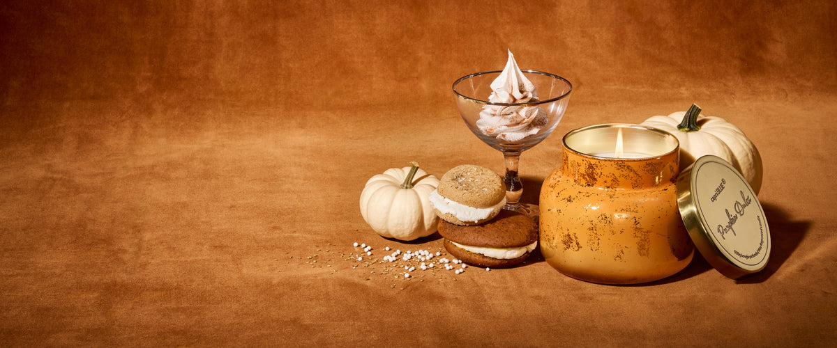 A lit, orange Pumpkin Dulce Glimmer candle beside whoopie pies, nonpareils, a dessert coupe with whipped cream and mini pumpkins.