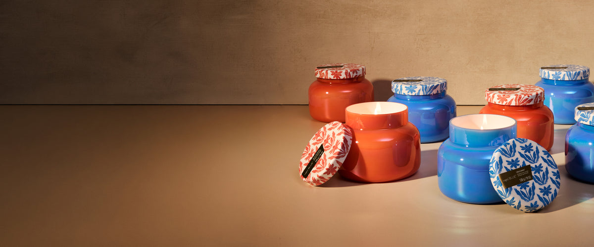 An lit, orange Isla Bloom Riviera Sunset candle beside a lit, blue Volcano Riviera Sunset candle arranged in front of several lidded Isla Bloom and Volcano candles.